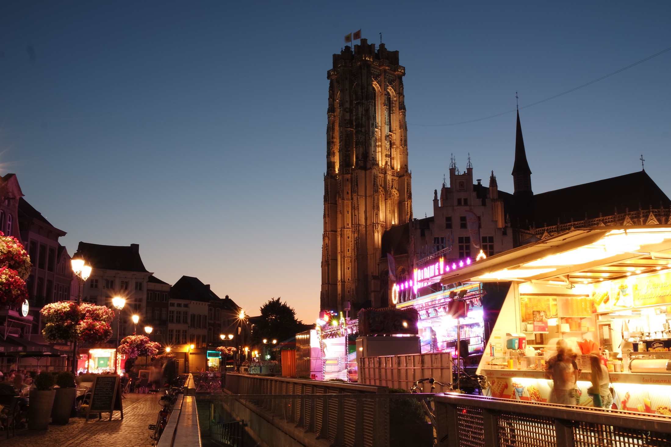 De verlichte Sint-Romboutstoren boven de avondlijke Grote Markt van Mechelen.