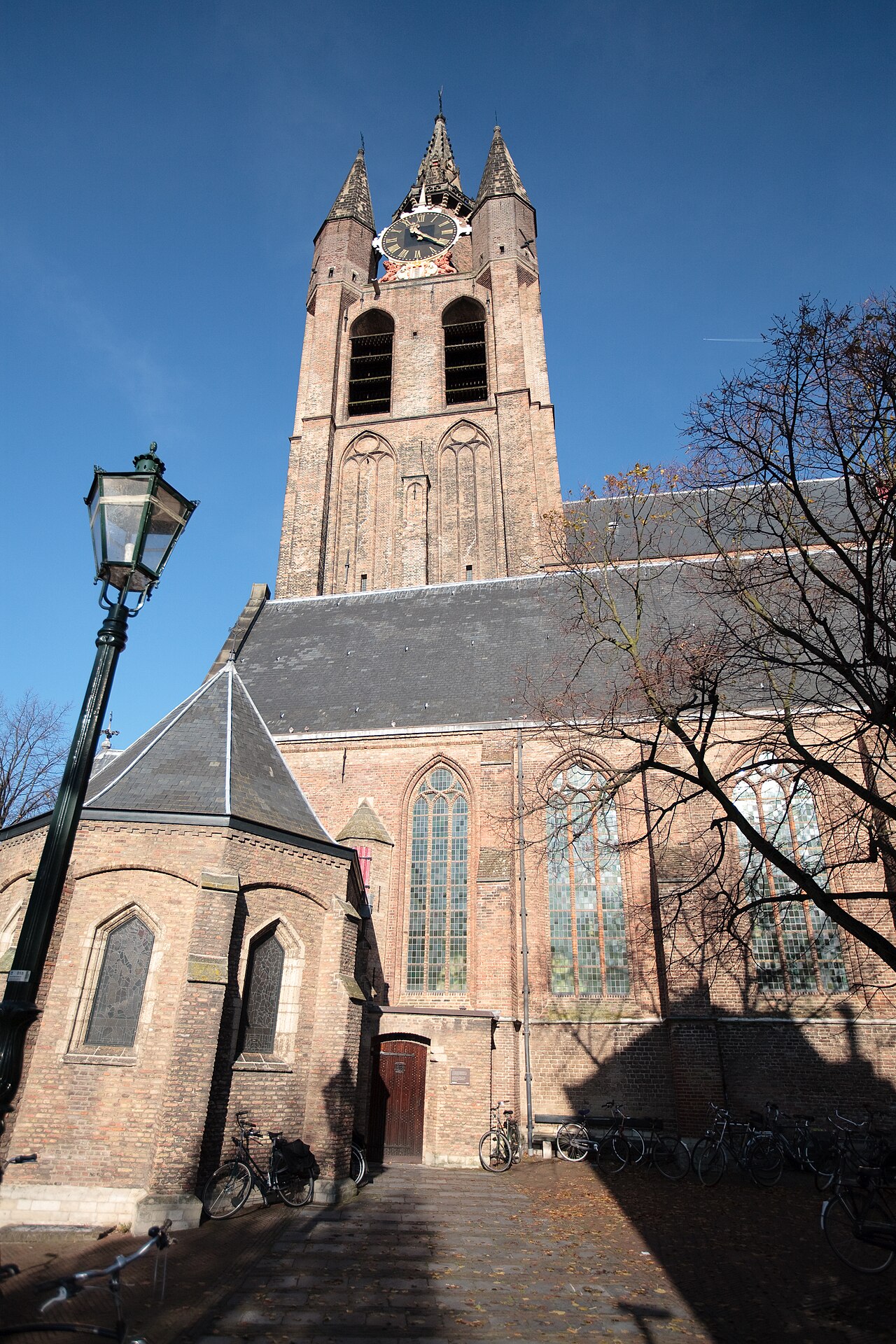 De bakstenen toren van de Oude Kerk in Delft tegen een blauwe lucht, met een lantaarn en bomen op de voorgrond.