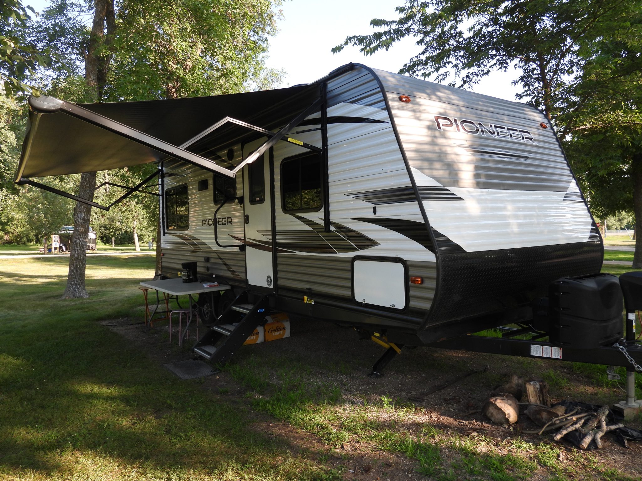 Een Pioneer-trekcaravan met uitgedraaide luifel en zichtbare dissel staat tussen bomen op een camping.