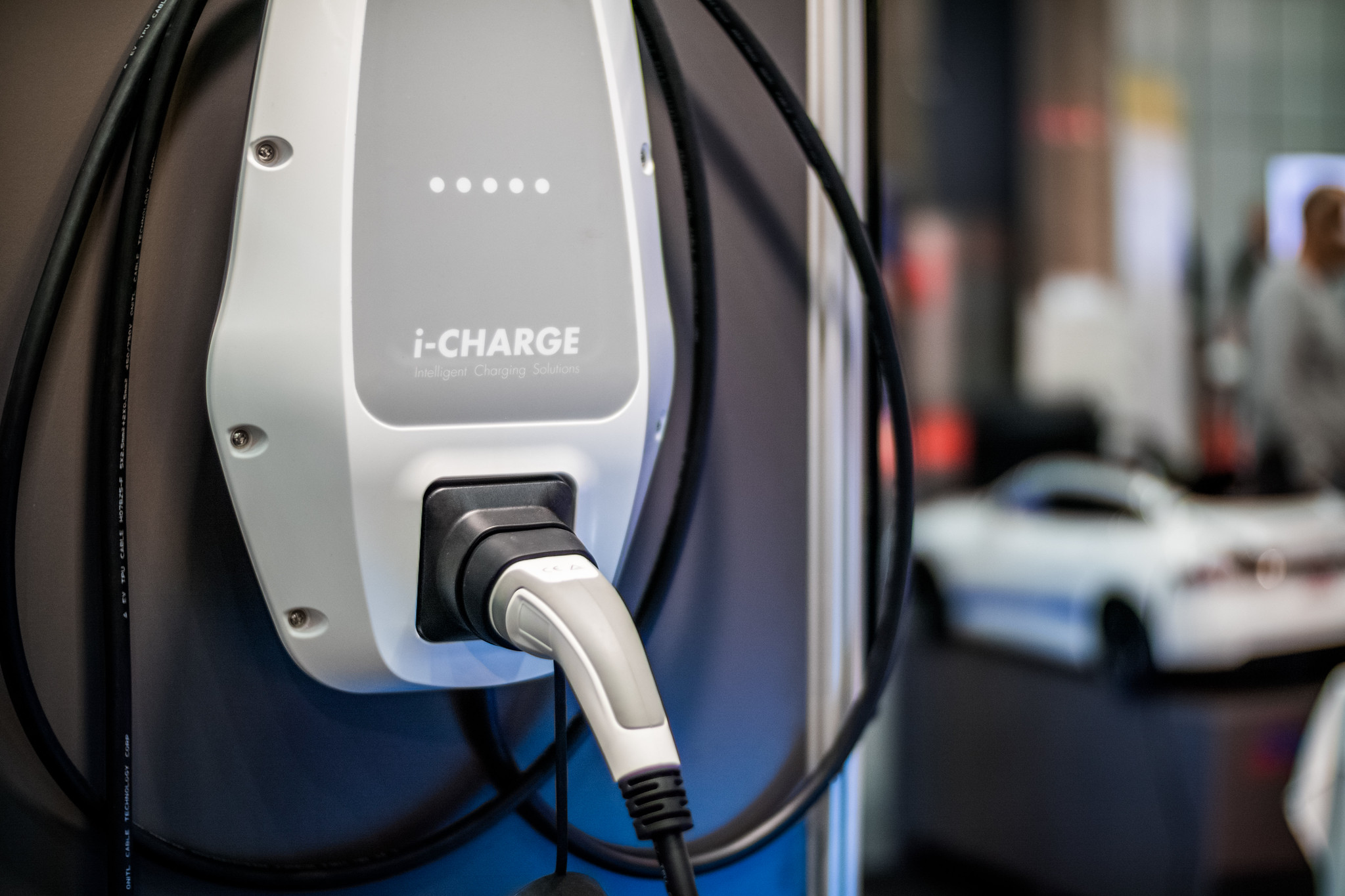 Close-up van i-CHARGE-laadapparatuur met een opgeborgen laadstekker en kabel; een auto staat onscherp op de achtergrond.