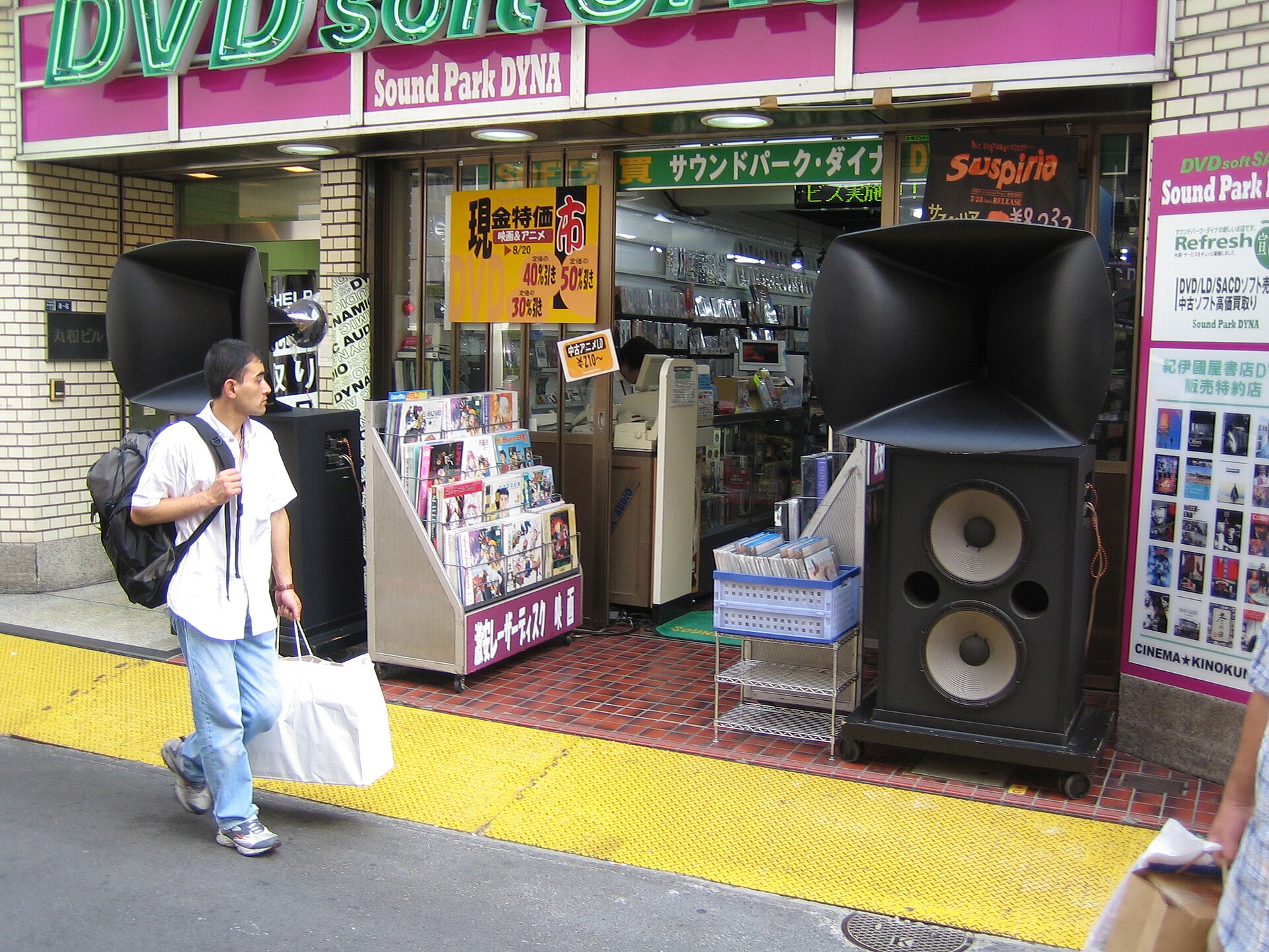 Twee grote luidsprekerkasten met ronde woofers en brede hoorns staan naast de ingang van een winkel in Tokio.