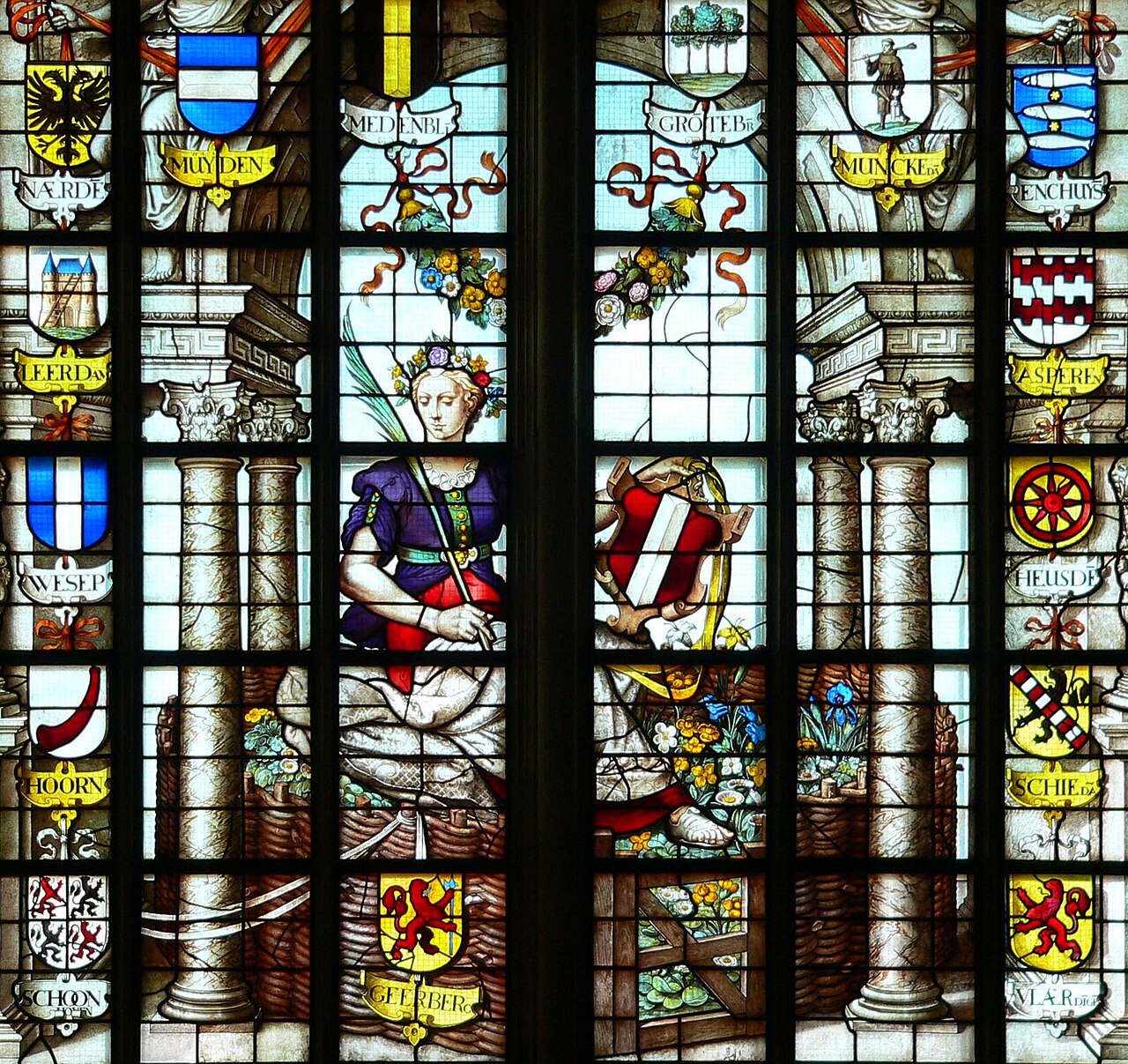 Detail van een kleurrijk gebrandschilderd raam met een zittende vrouwenfiguur, stadswapens en zuilen.