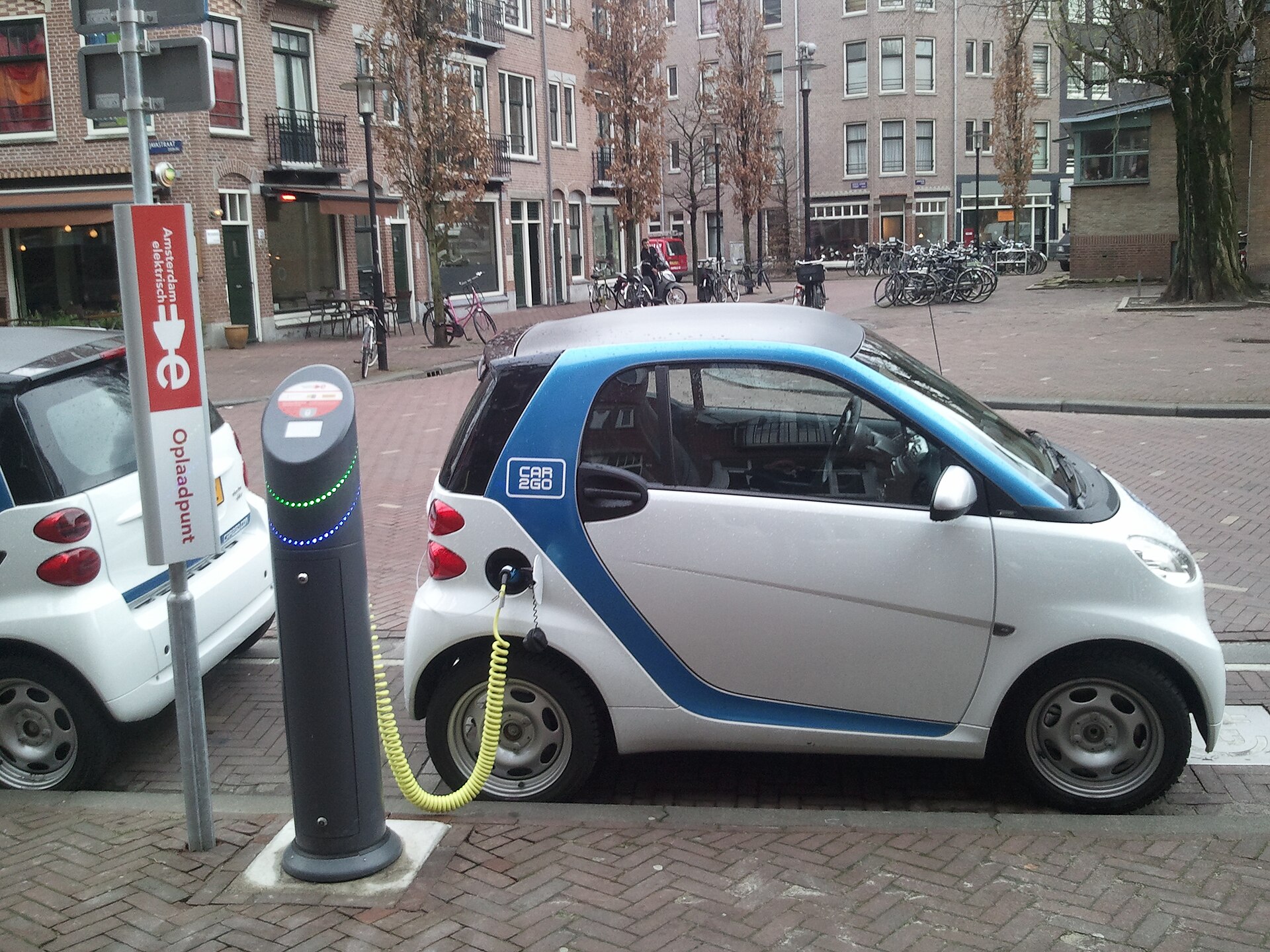 Een witte en blauwe elektrische Smart staat met een groene kabel aan een laadpaal bij Amsterdamse grachtenpanden.