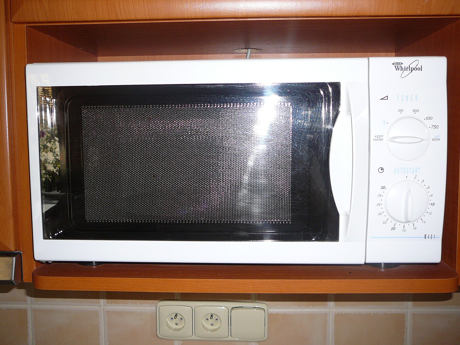 Een witte magnetron met een donkere deur en twee draaiknoppen staat in een houten nis.