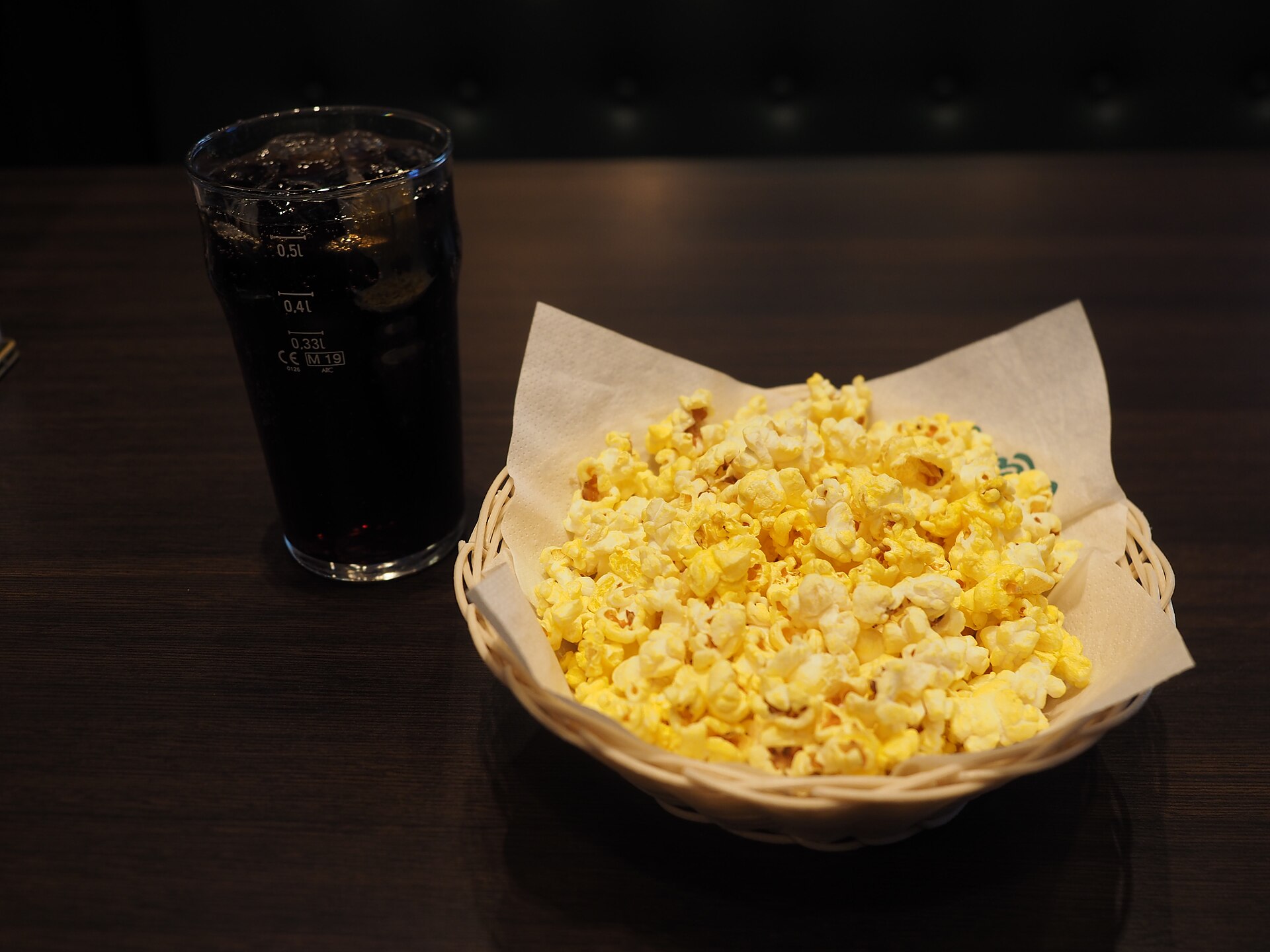 Een kom gele popcorn staat naast een glas frisdrank op een donkere tafel.