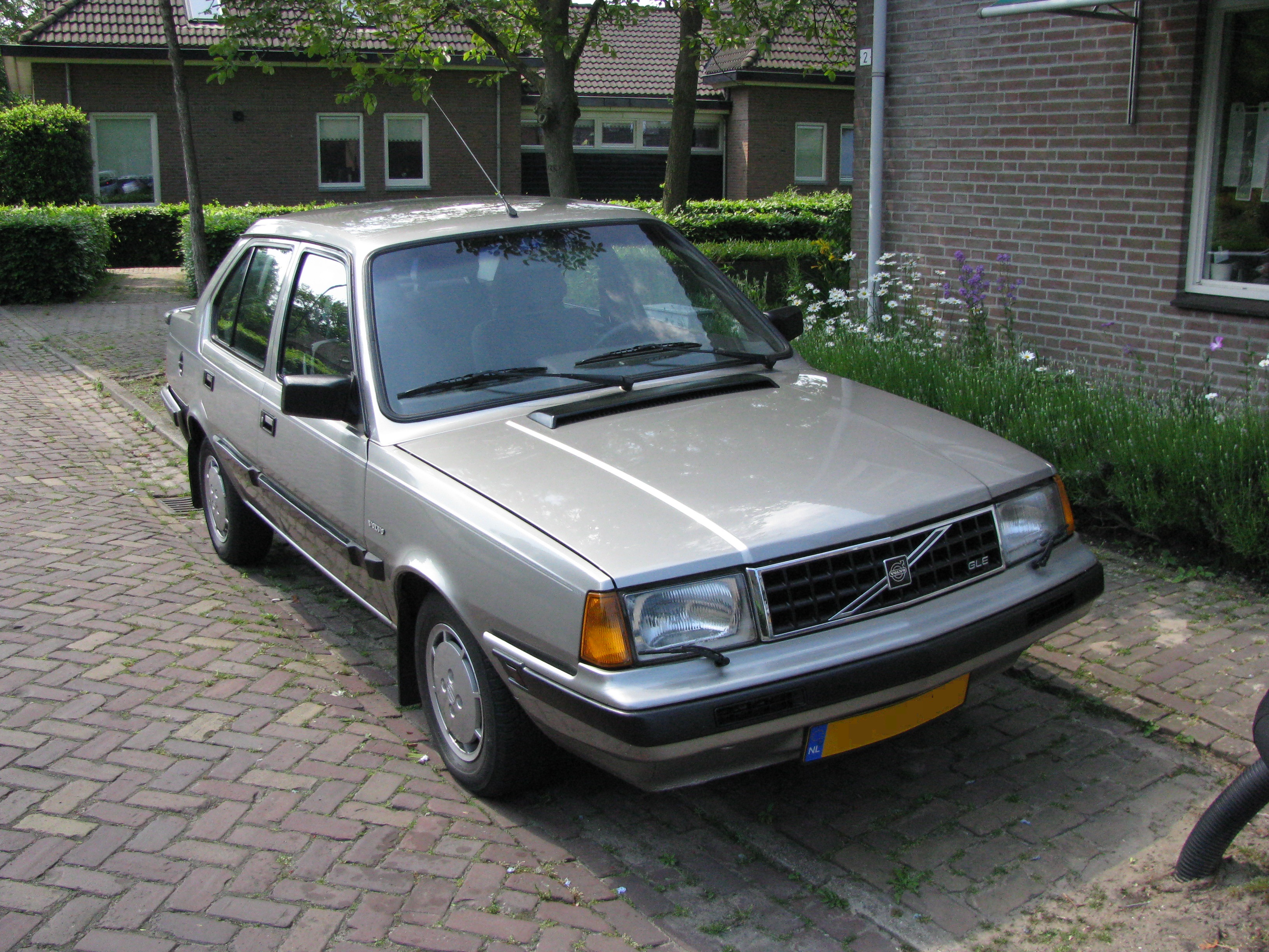 Een zilvergrijze vierdeurs Volvo 340 GLE met Nederlands kenteken, geparkeerd voor een woning.